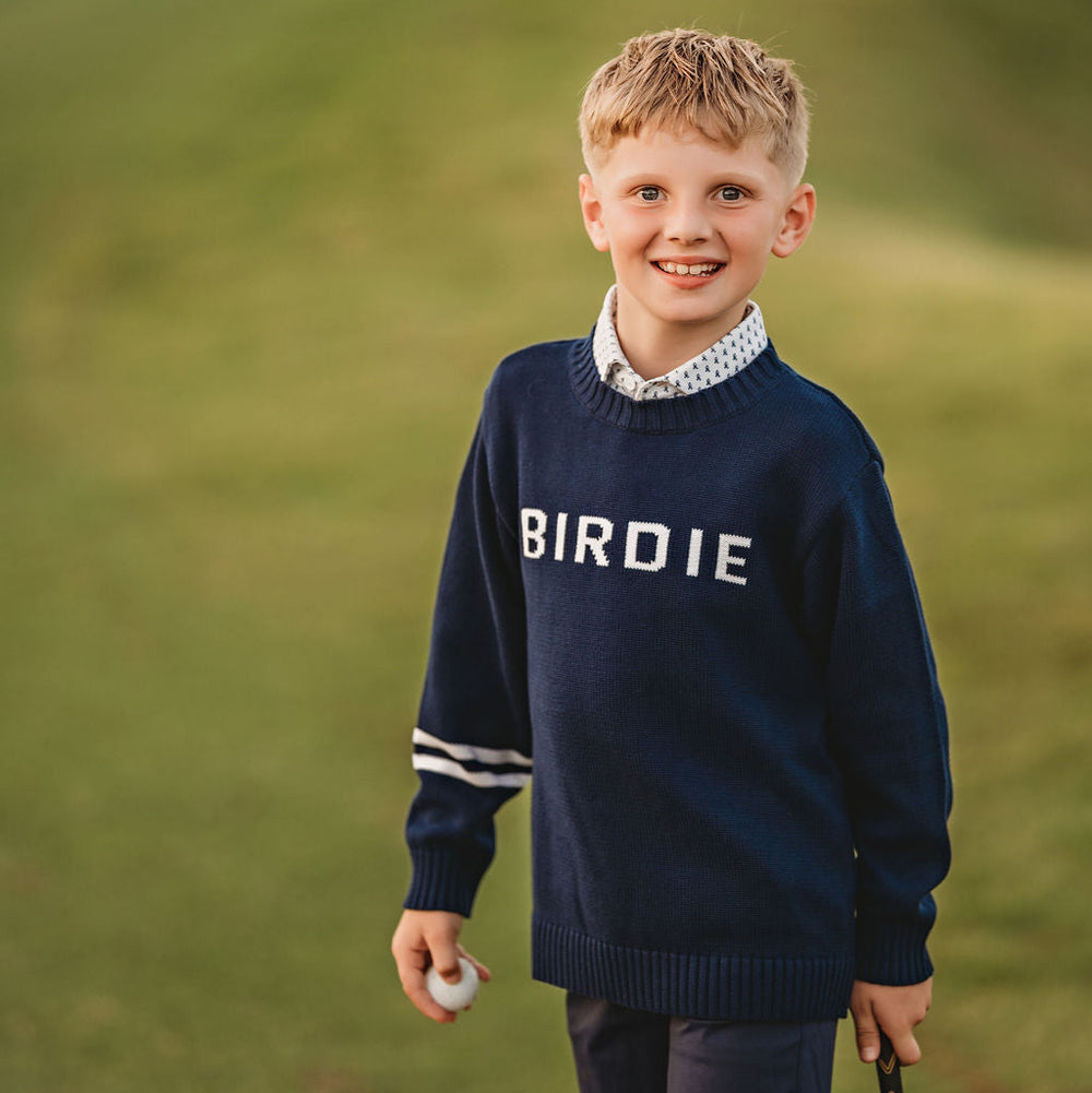 Young boy in a navy sweater with 'BIRDIE' on it standing on a golf course.